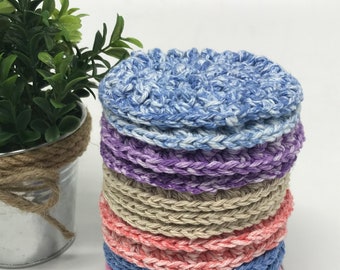 May include: A stack of handmade, round coasters in various colors, including blue, purple, tan, pink, and gray. The coasters are made of a textured, crocheted material. A small potted plant is visible in the background.