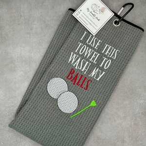 May include: Gray golf towel with the text "I USE THIS TOWEL TO WASH MY BALLS" in white and red. The towel features embroidered golf balls and a tee. A black carabiner clip is attached.