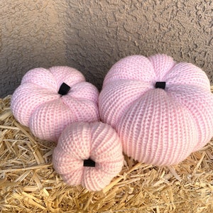 May include: Four pink knitted pumpkins of varying sizes are arranged on a bed of straw. Each pumpkin has a ribbed texture and a small black stem. The pumpkins are a soft, pastel pink color.