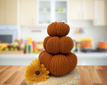 May include: A stack of three knitted orange pumpkins of varying sizes, arranged on a decorative wooden tray. A yellow and orange artificial flower is placed on the tray. The pumpkins are set against a blurred kitchen background.