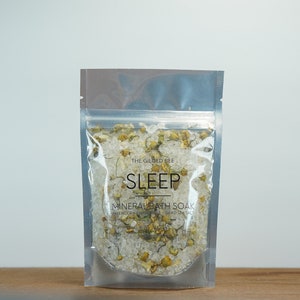 May include: A silver pouch filled with white bath salts and dried chamomile flowers. The pouch has a label that reads "The Gilded Bee" and "SLEEP MINERAL BATH SOAK".