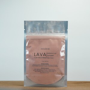 May include: A clear plastic pouch containing a pink clay face mask powder. The pouch has a label that reads "The Gilded Bee Lava Clay Face Mask" with the ingredients listed as "Rhassoul Clay, Acai Berry, Beet Root".