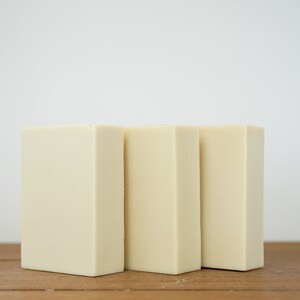 May include: Three rectangular bars of ivory-coloured soap arranged in a row on a wooden surface.
