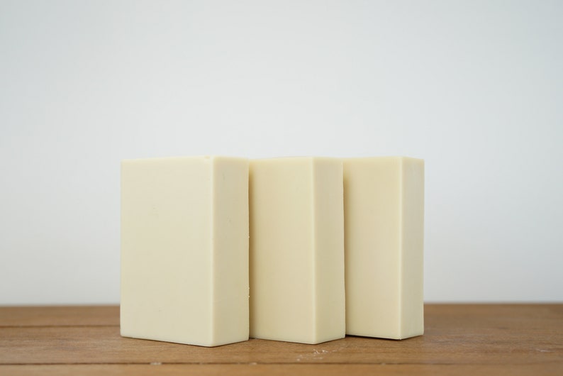May include: Three rectangular bars of ivory-coloured soap arranged in a row on a wooden surface.
