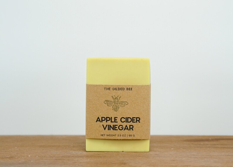 May include: A yellow bar of soap with a brown label that reads "The Gilded Bee" and "Apple Cider Vinegar" with a bee illustration. The label also states "Net Weight 3.5 oz | 99 g".