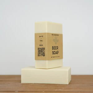 May include: Two bars of white soap stacked on top of each other. The top bar has a brown paper label that reads "Save the Bees" and "The Salted Bee Beer Soap" with a bee illustration and a QR code. The net weight is 100g (3.5 oz).