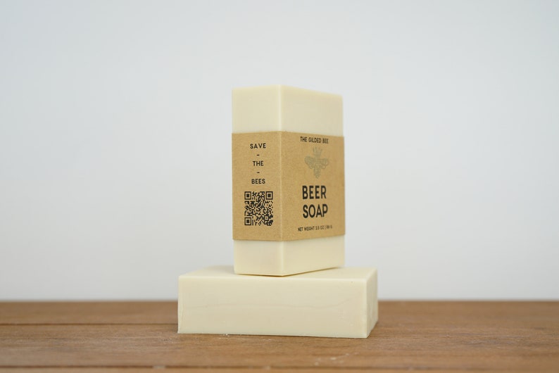 May include: Two bars of white soap stacked on top of each other. The top bar has a brown paper label that reads "Save the Bees" and "The Salted Bee Beer Soap" with a bee illustration and a QR code. The net weight is 100g (3.5 oz).