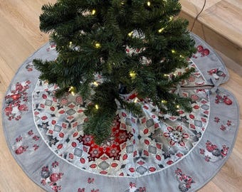 Puede incluir: Un pequeño árbol de Navidad artificial con luces blancas cálidas. El árbol se encuentra sobre una falda de árbol gris con un patrón festivo de adornos rojos y plateados. El árbol está sobre un suelo de madera.