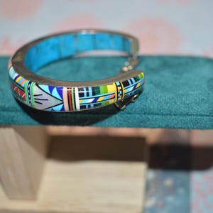 May include: A silver-toned cuff bracelet with a turquoise inlay and colorful geometric patterns. The bracelet features a hinged design and is displayed on a teal velvet surface.