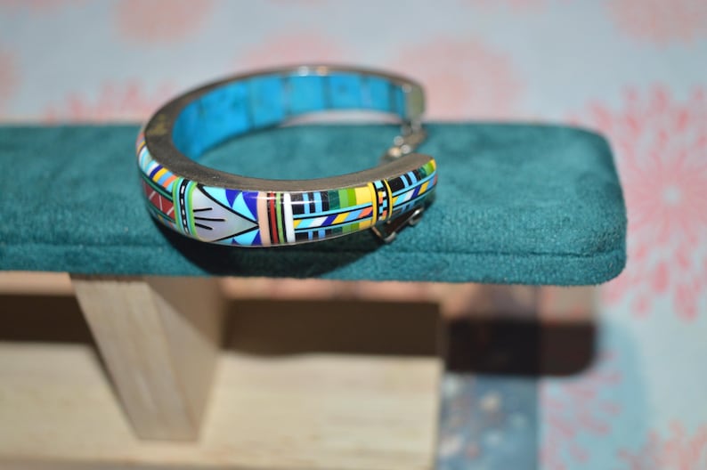 May include: A silver-toned cuff bracelet with a turquoise inlay and colorful geometric patterns. The bracelet features a hinged design and is displayed on a teal velvet surface.