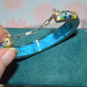 May include: A silver bracelet with a turquoise inlay and colorful mosaic accents. The bracelet has a hinged design with a chain closure. The mosaic features blue, yellow, green, and red colors.