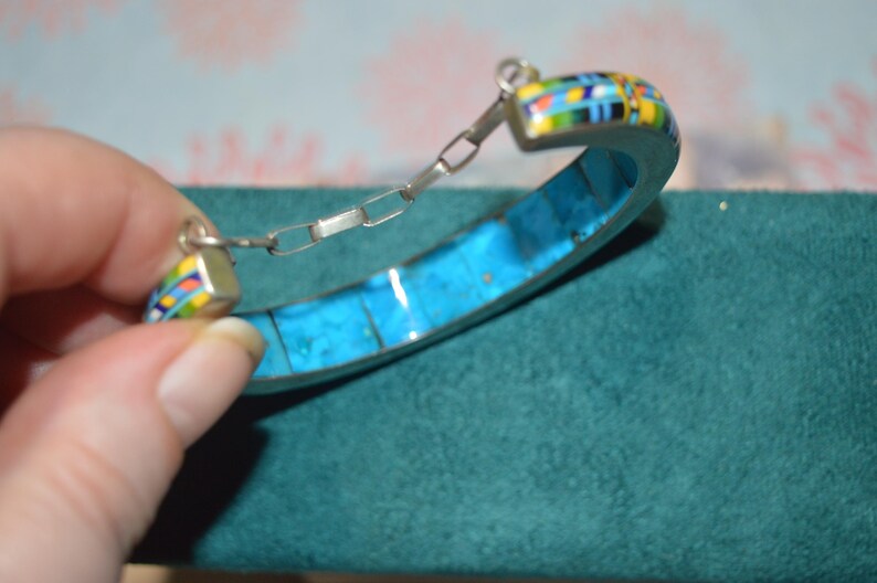 May include: A silver bracelet with a turquoise inlay and colorful mosaic accents. The bracelet has a hinged design with a chain closure. The mosaic features blue, yellow, green, and red colors.