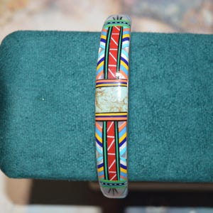 May include: A colorful bracelet with a geometric design. The bracelet features a central section of light green stone, bordered by vibrant patterns of red, blue, yellow, and white. The bracelet is displayed on a teal surface.
