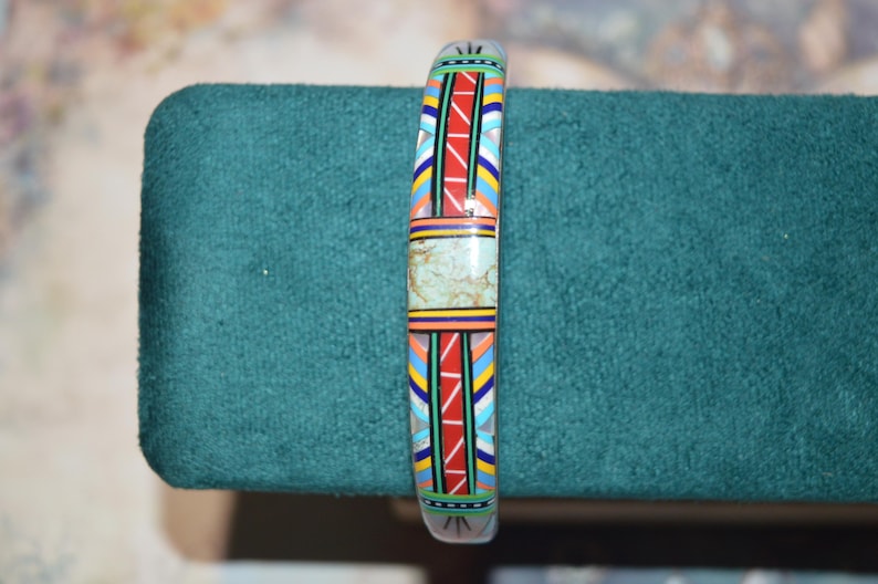 May include: A colorful bracelet with a geometric design. The bracelet features a central section of light green stone, bordered by vibrant patterns of red, blue, yellow, and white. The bracelet is displayed on a teal surface.