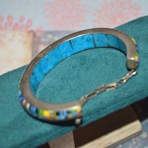May include: Silver-toned bracelet with a turquoise inlay and colorful mosaic accents. The bracelet features a hinged design with a chain closure. The turquoise is a bright blue color. The mosaic includes yellow, blue, and white. The bracelet is displayed on a teal surface.