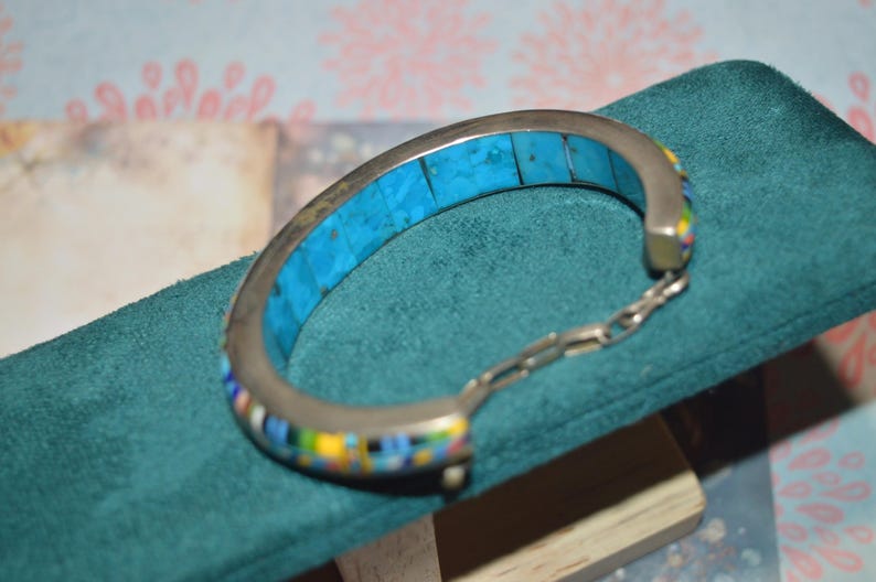 May include: Silver-toned bracelet with a turquoise inlay and colorful mosaic accents. The bracelet features a hinged design with a chain closure. The turquoise is a bright blue color. The mosaic includes yellow, blue, and white. The bracelet is displayed on a teal surface.