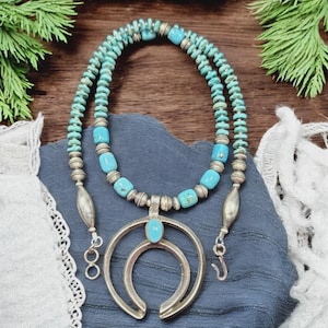 Puede incluir: Un collar turquesa y plateado con un gran colgante en forma de media luna. El collar presenta dos hilos de pequeñas cuentas turquesas y cuentas turquesas más grandes. El colgante es plateado con una piedra turquesa en el centro.