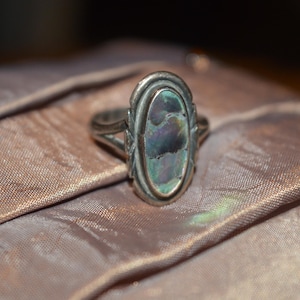 May include: A silver ring with an oval, iridescent abalone shell center stone. The ring has a decorative border and is set on a light brown, textured fabric. The ring's design is vintage and elegant.