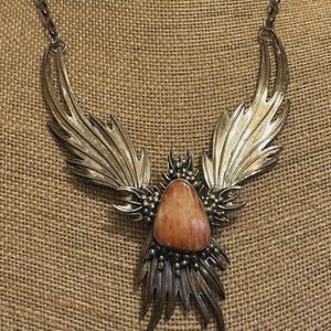 May include: A silver-toned statement necklace with a central, orange-toned stone. The necklace features stylized wing-like elements and intricate detailing, creating a bold, artistic design. The chain is a simple, linked design.