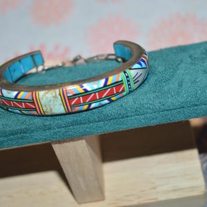 May include: A colorful cuff bracelet with a mosaic design. The bracelet features a silver-tone metal band and is adorned with turquoise, red, yellow, and white geometric patterns. The bracelet is displayed on a teal velvet surface.