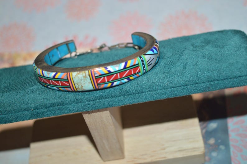 May include: A colorful cuff bracelet with a mosaic design. The bracelet features a silver-tone metal band and is adorned with turquoise, red, yellow, and white geometric patterns. The bracelet is displayed on a teal velvet surface.