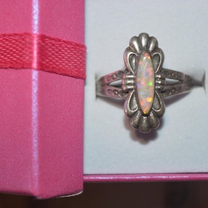 May include: A silver ring with an oval opal gemstone, set in a decorative floral design. The ring is displayed in a pink gift box with a red ribbon. The opal has a play of color.