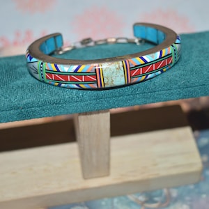 May include: A silver-toned cuff bracelet with a colorful geometric design. The bracelet features turquoise accents and a central turquoise stone. The design includes red, yellow, blue, and white patterns. The bracelet is displayed on a teal surface.
