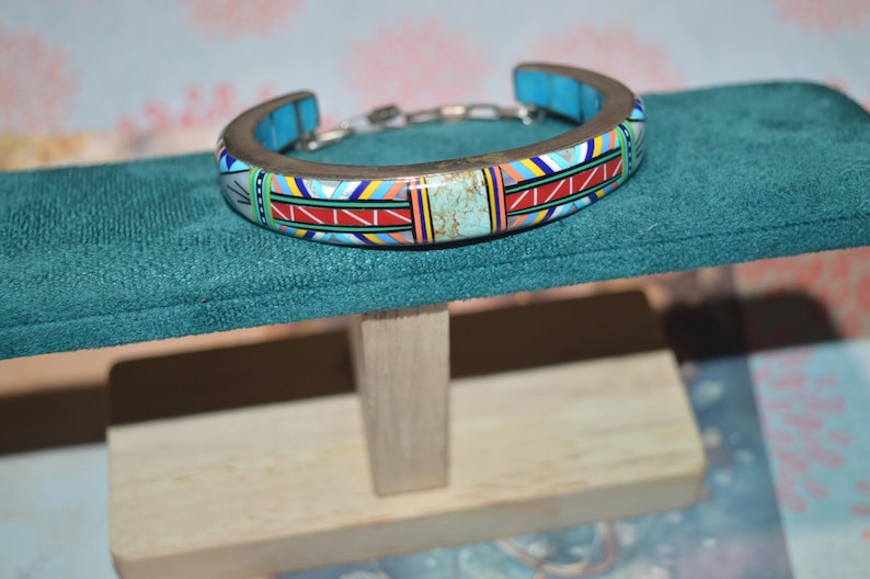 May include: A silver-toned cuff bracelet with a colorful geometric design. The bracelet features turquoise accents and a central turquoise stone. The design includes red, yellow, blue, and white patterns. The bracelet is displayed on a teal surface.