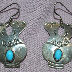 May include: A pair of silver earrings featuring a turquoise stone. The earrings are vase-shaped with decorative patterns and a hook. The background is a muted purple fabric. The earrings are made of silver.