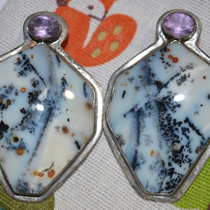 Puede incluir: Un par de pendientes plateados. Cada pendiente presenta una piedra de ágata dendrítica con base blanca, inclusiones negras y marrones, y una amatista en la parte superior. Los pendientes tienen forma geométrica.