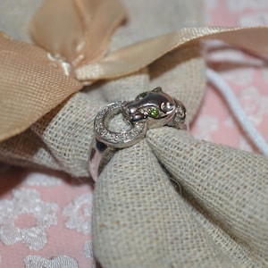 May include: A silver ring featuring a panther head with green eyes and a circular design with small, clear stones. The ring is set against a beige fabric and a pink floral background.