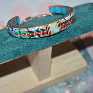 May include: A colorful cuff bracelet with a mosaic design, featuring turquoise, red, yellow, and white geometric patterns. The bracelet is displayed on a teal green velvet surface, showcasing its intricate details and craftsmanship. The bracelet is made of metal and has a chain link closure.