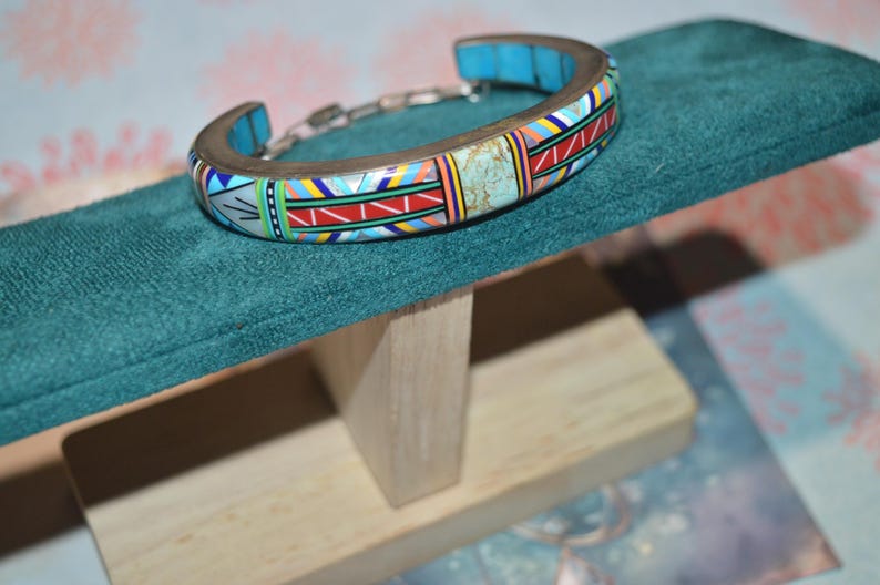 May include: A colorful cuff bracelet with a mosaic design, featuring turquoise, red, yellow, and white geometric patterns. The bracelet is displayed on a teal green velvet surface, showcasing its intricate details and craftsmanship. The bracelet is made of metal and has a chain link closure.