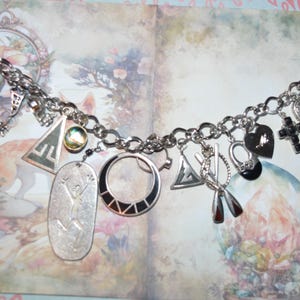 Pode incluir: Pulseira de berloques prateada com vários berloques, incluindo uma cruz, um coração e formas geométricas. A pulseira é exibida em um fundo de uma ilustração floral e de raposa, criando uma estética caprichosa. Os berloques variam em tamanho e design.
