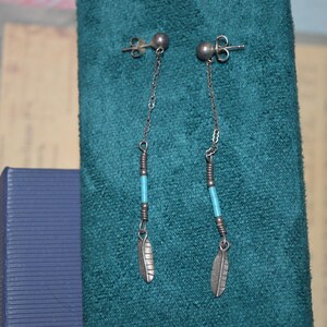 May include: A pair of silver-toned dangle earrings. Each earring features a small ball stud, a delicate chain, a turquoise bead, and a feather charm. The earrings are displayed against a teal background.