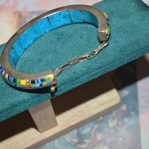 May include: Silver-tone hinged bracelet with a turquoise inlay and colorful mosaic accents. The bracelet is displayed on a teal green velvet surface, showcasing its intricate design and craftsmanship. A stylish piece of jewelry.
