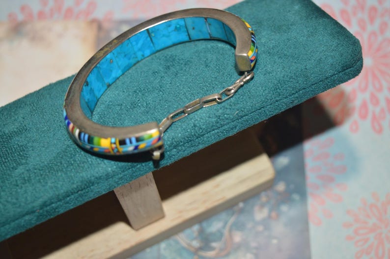 May include: Silver-tone hinged bracelet with a turquoise inlay and colorful mosaic accents. The bracelet is displayed on a teal green velvet surface, showcasing its intricate design and craftsmanship. A stylish piece of jewelry.
