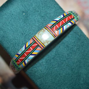 May include: A silver-toned bracelet with a colorful geometric pattern. The design includes red, blue, yellow, and green accents, with a central turquoise-colored stone. The bracelet is displayed on a teal green surface.