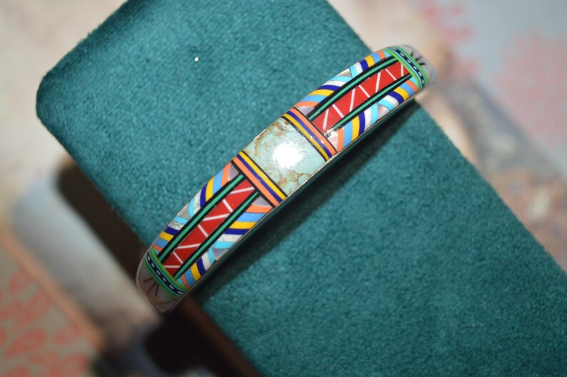 May include: A silver-toned bracelet with a colorful geometric pattern. The design includes red, blue, yellow, and green accents, with a central turquoise-colored stone. The bracelet is displayed on a teal green surface.