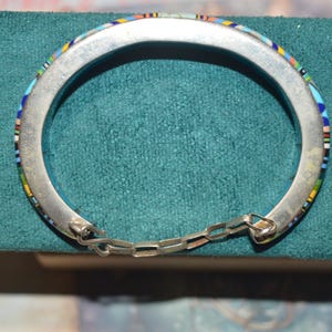 May include: Silver-tone hinged bracelet with a mosaic inlay of colorful stones. The bracelet features a chain closure and is displayed on a teal surface. The mosaic design includes blue, yellow, red, and white elements.