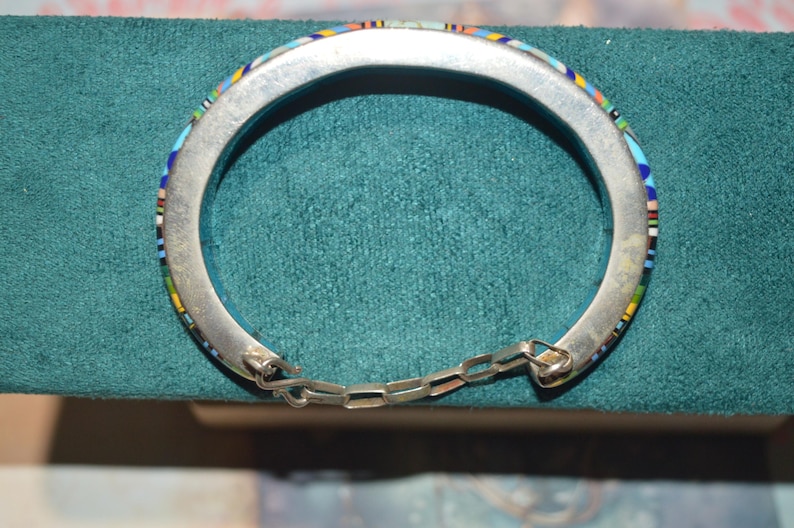 May include: Silver-tone hinged bracelet with a mosaic inlay of colorful stones. The bracelet features a chain closure and is displayed on a teal surface. The mosaic design includes blue, yellow, red, and white elements.
