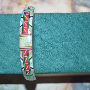 May include: A colorful bracelet with a geometric pattern in red, blue, yellow, and white. The bracelet has a central rectangular section with a light green, speckled design. The bracelet is displayed on a teal green surface.
