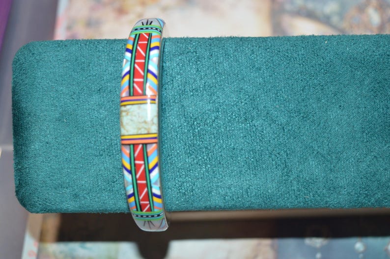May include: A colorful bracelet with a geometric pattern in red, blue, yellow, and white. The bracelet has a central rectangular section with a light green, speckled design. The bracelet is displayed on a teal green surface.