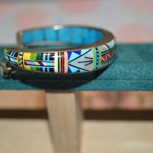 May include: A silver-toned cuff bracelet with a vibrant, geometric design. The bracelet features a turquoise interior and is adorned with colorful patterns in yellow, blue, red, and green. The design is reminiscent of Native American art.