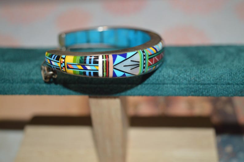 May include: A silver-toned cuff bracelet with a vibrant, geometric design. The bracelet features a turquoise interior and is adorned with colorful patterns in yellow, blue, red, and green. The design is reminiscent of Native American art.