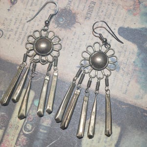 May include: A pair of silver-toned dangle earrings. Each earring has a floral design with a central circular element and dangling rectangular and teardrop-shaped pendants. The earrings are on a vintage paper background.