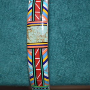 May include: A colorful bracelet with a central turquoise-colored stone. The bracelet features a geometric pattern with red, blue, yellow, and white accents. The design is reminiscent of Native American art.