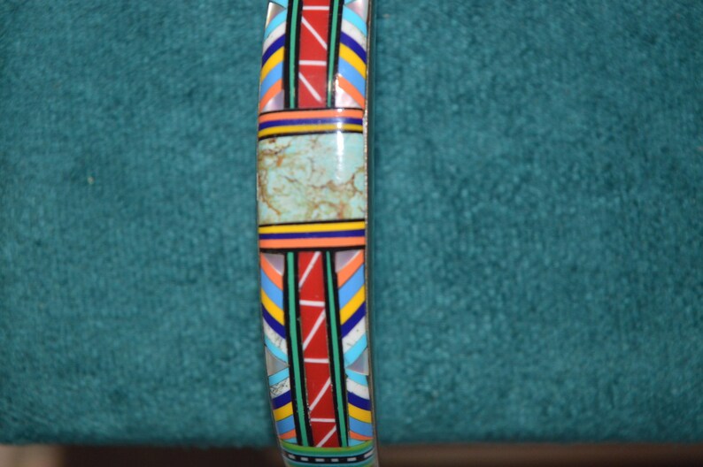 May include: A colorful bracelet with a central turquoise-colored stone. The bracelet features a geometric pattern with red, blue, yellow, and white accents. The design is reminiscent of Native American art.