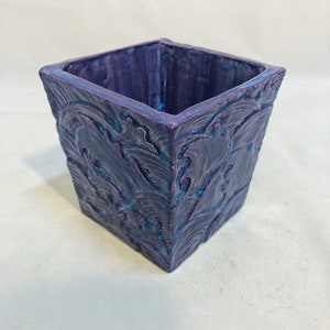 May include: A square purple planter with a textured, wave-like design. The planter has a dark purple base with teal accents. Suitable for indoor or outdoor use. The planter's dimensions are approximately 15 cm x 15 cm.