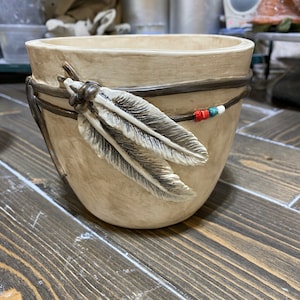 Op de afbeelding: Een beige keramische plantenbak met een decoratief veermotief. De plantenbak is versierd met een donkerbruine band en veren, en kleine rode, witte en blauwe kralen. De plantenbak staat op een houten oppervlak.